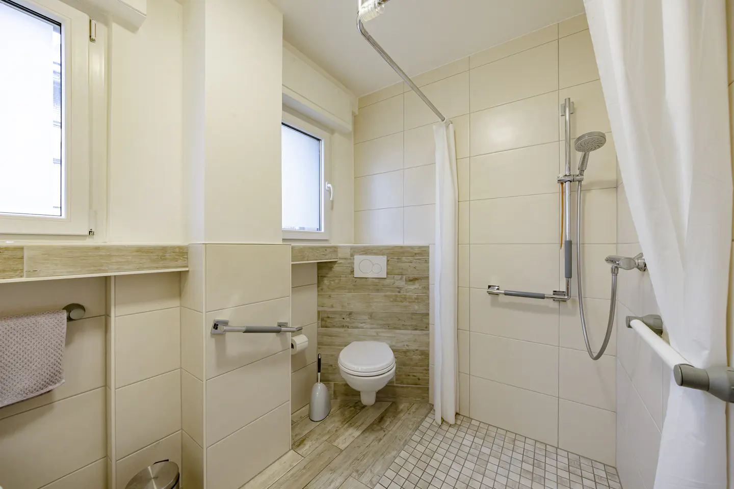 Bright bathroom with beige tiles, white toilet, and shower with grab bars and a white curtain. Two windows provide natural light.