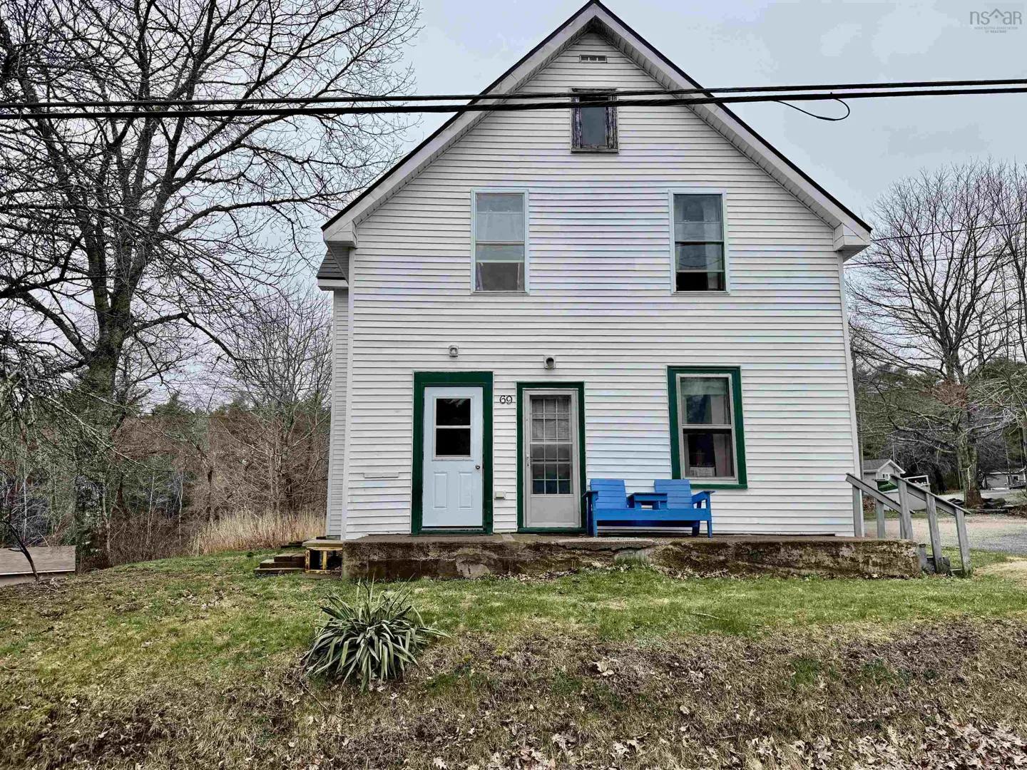 Two-story white house with green trim, two doors, and a blue bench on the porch. Bare trees surround the house.