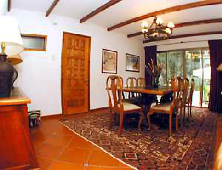 A dining room with a long wooden table and chairs on a patterned rug. A chandelier hangs from the ceiling with exposed wooden beams. A carved wooden door is on the left.