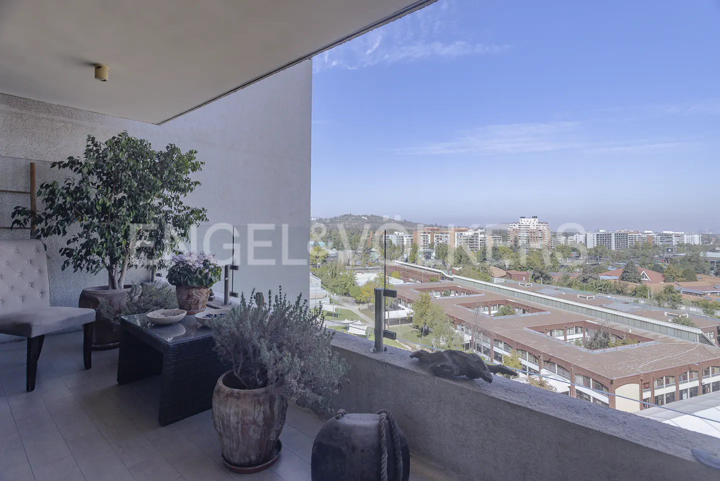 Balcony view with potted plants, chair, and table overlooking a city skyline under a blue sky.