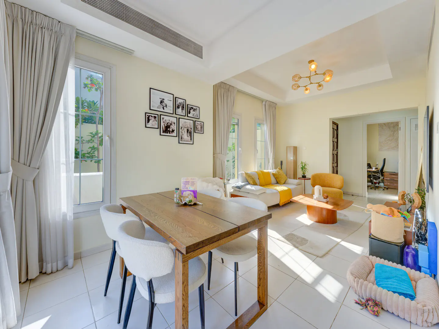 A bright, open-plan living and dining area with a wooden table, white chairs, and a white sofa with yellow accents. Black and white photos adorn the wall.