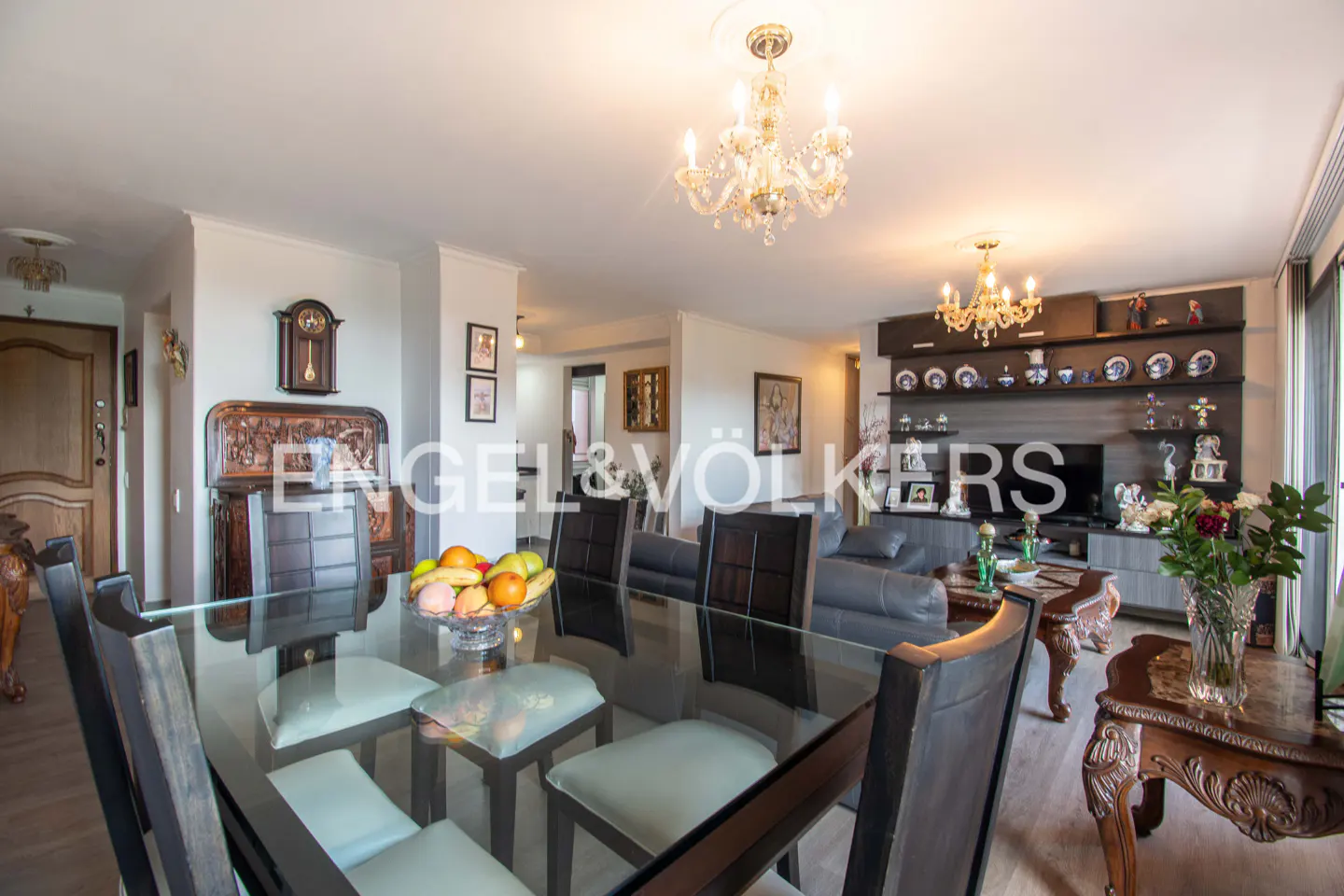 A bright, open-concept living and dining area with a glass-top table, chandelier, and decorative shelves.