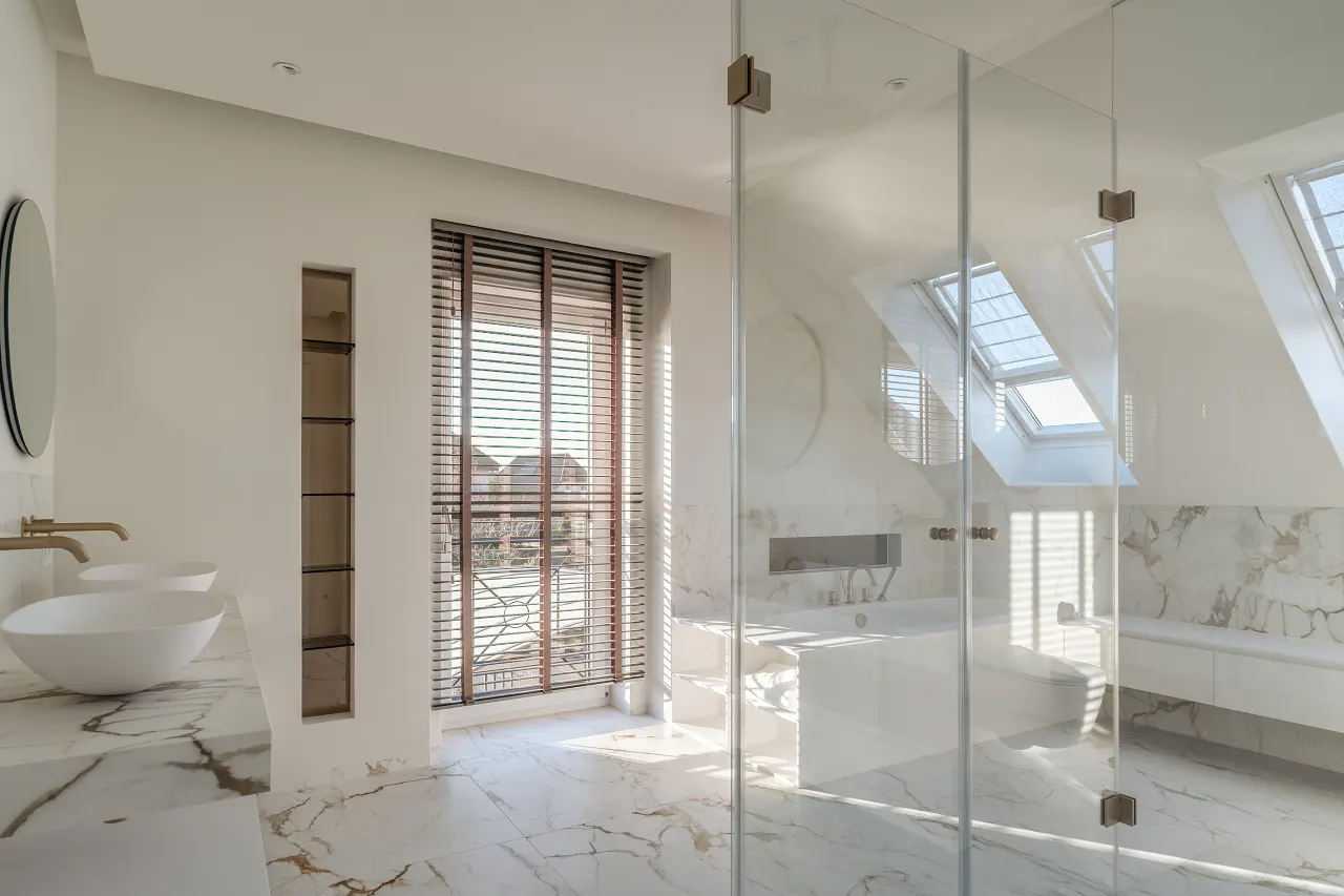 Bright bathroom with marble floors, white walls, and a glass shower. Two white sinks sit on a marble countertop. A window with blinds lets in sunlight.