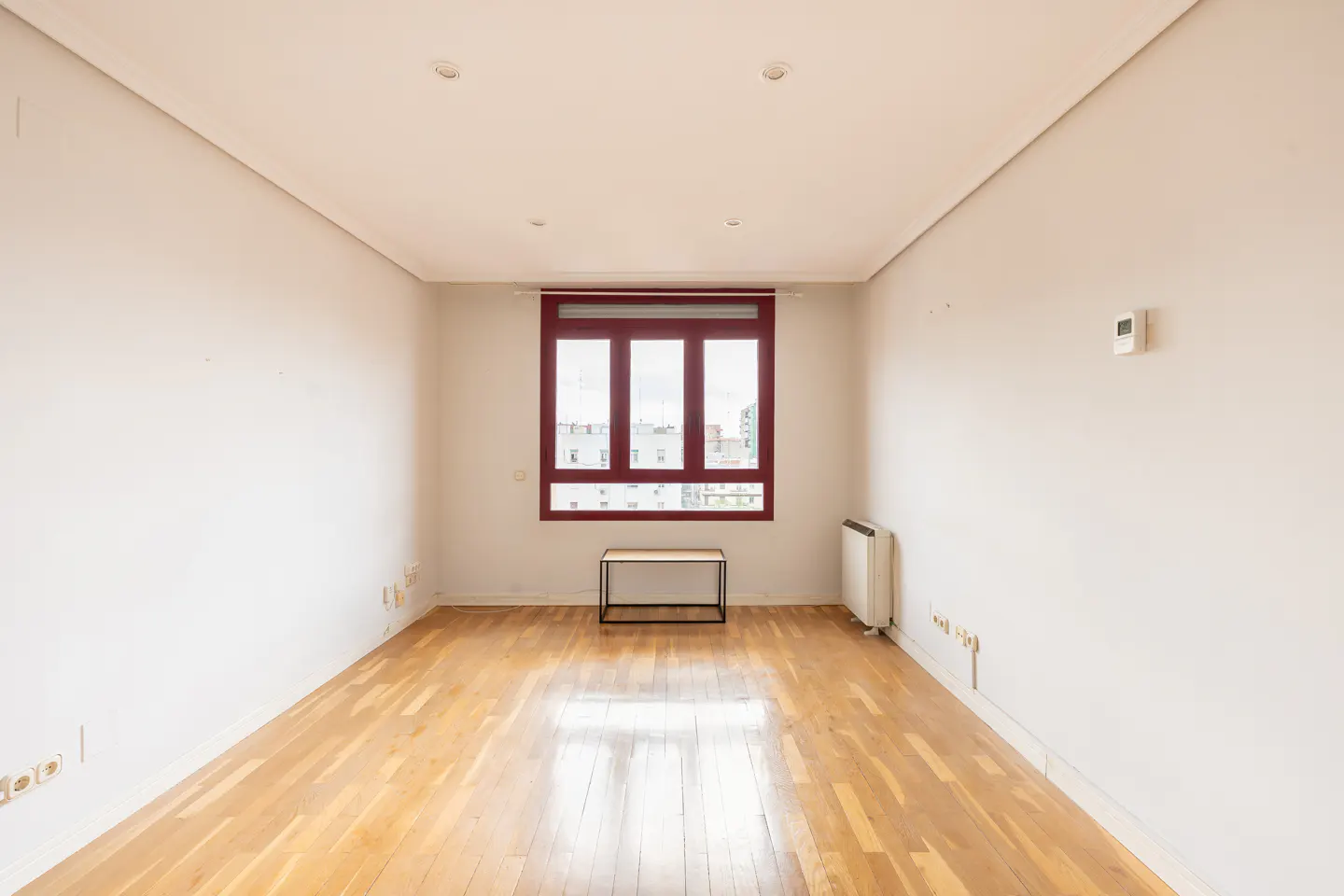 Empty room with light wood floors, white walls, and a red-framed window. A small table sits beneath the window.