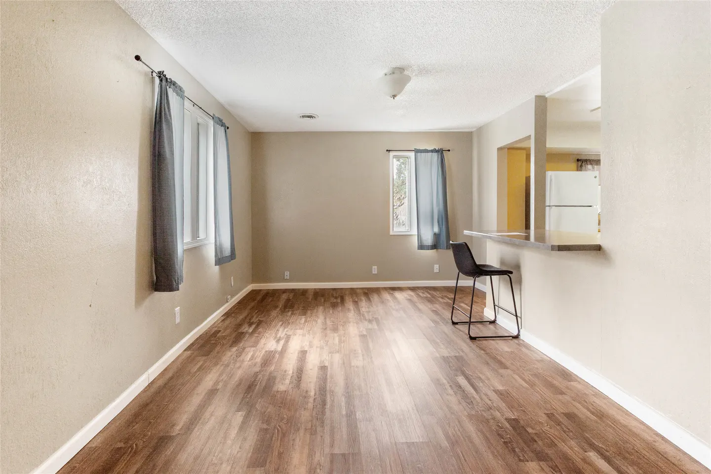 A bright, empty living room with wood floors and beige walls. A black bar stool sits near a kitchen counter. Curtains hang on windows.