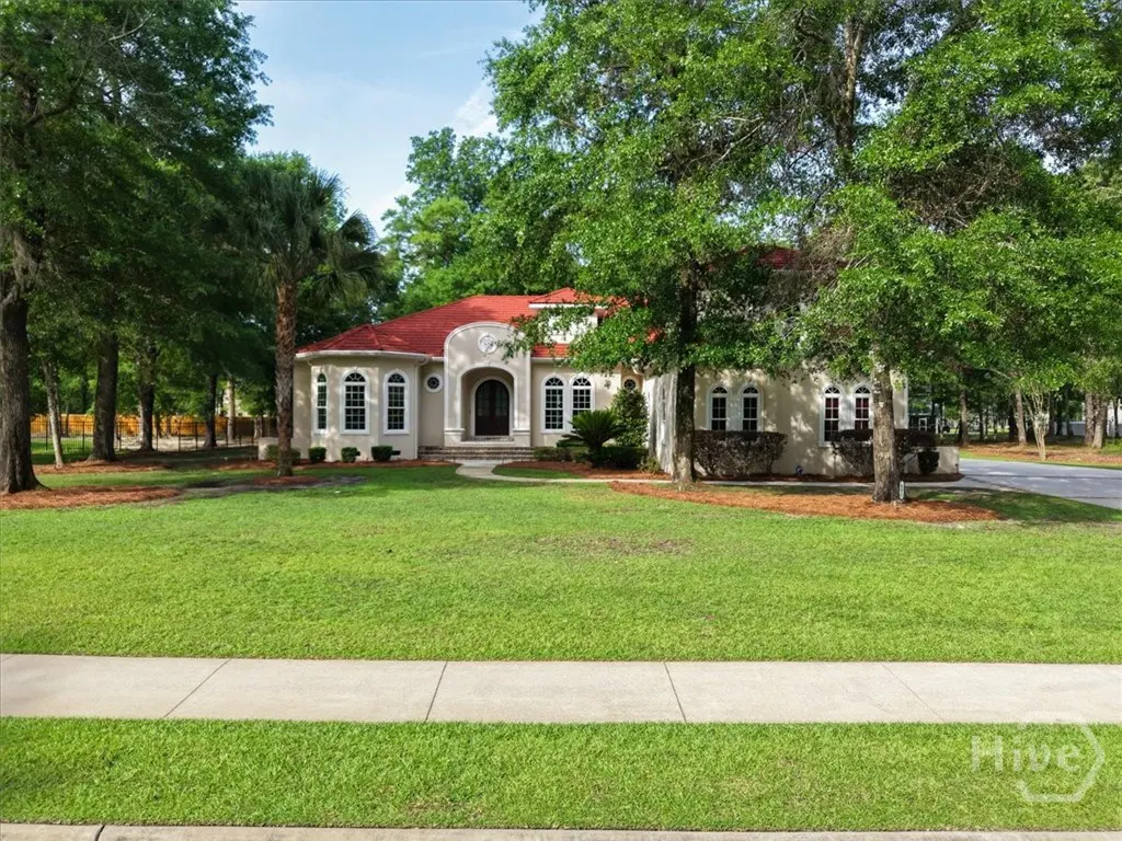 Beige house with a red roof, surrounded by green trees and a lawn. A sidewalk is in the foreground.