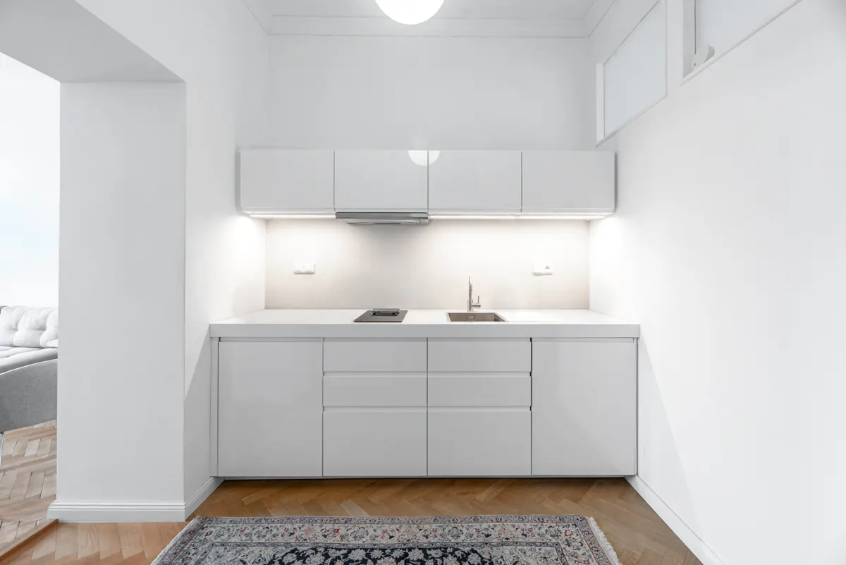 Bright, modern kitchen with white cabinets, countertops, and walls. A patterned rug sits on a wood floor.