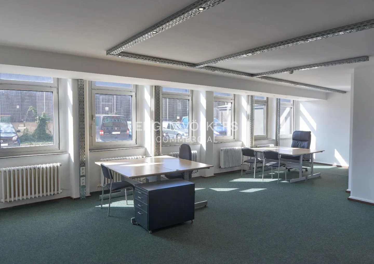 Bright office space with green carpet, desks, chairs, and large windows. Radiators are visible under the windows.