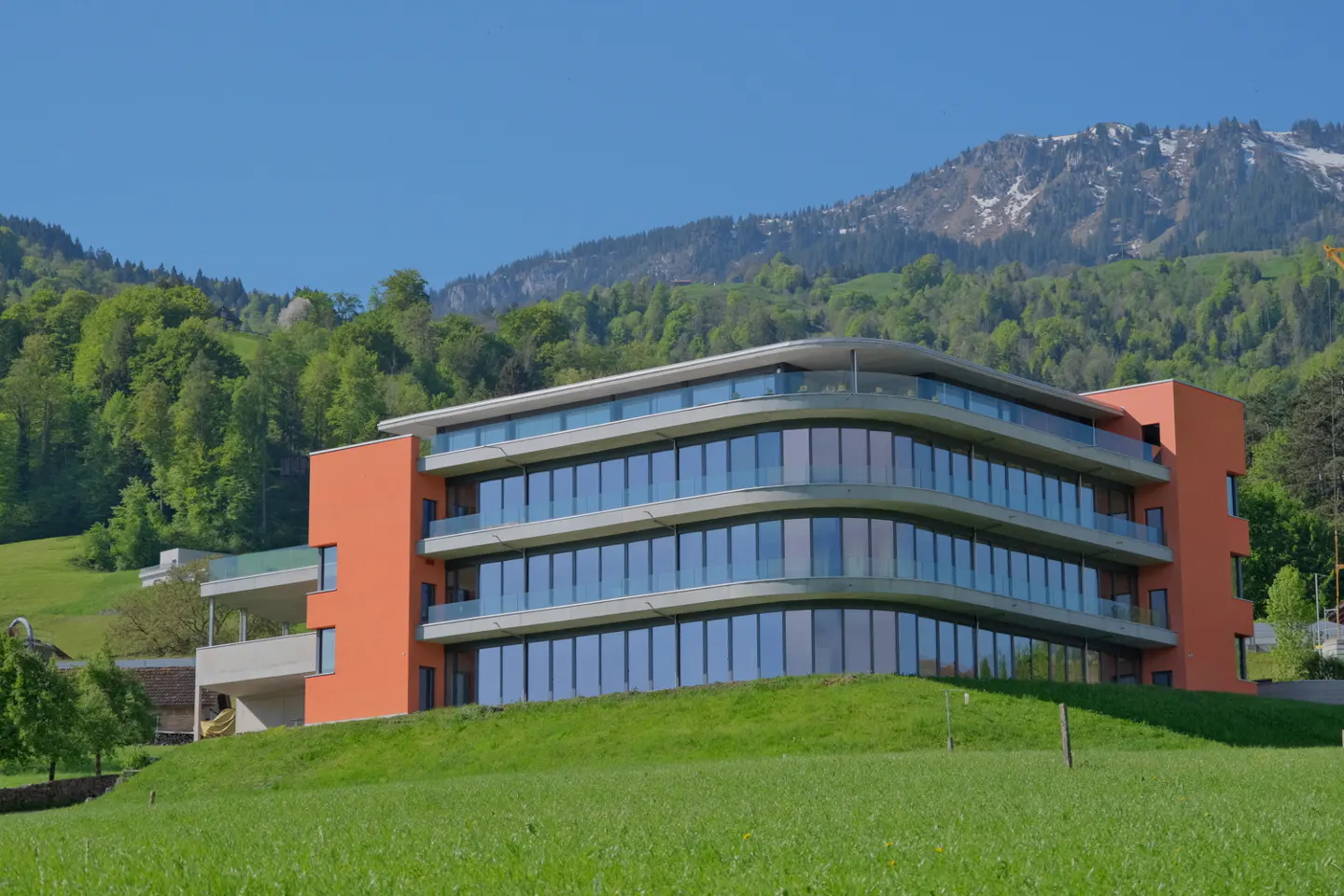 Modern orange building with curved glass windows on a green hill, mountains and blue sky in the background.