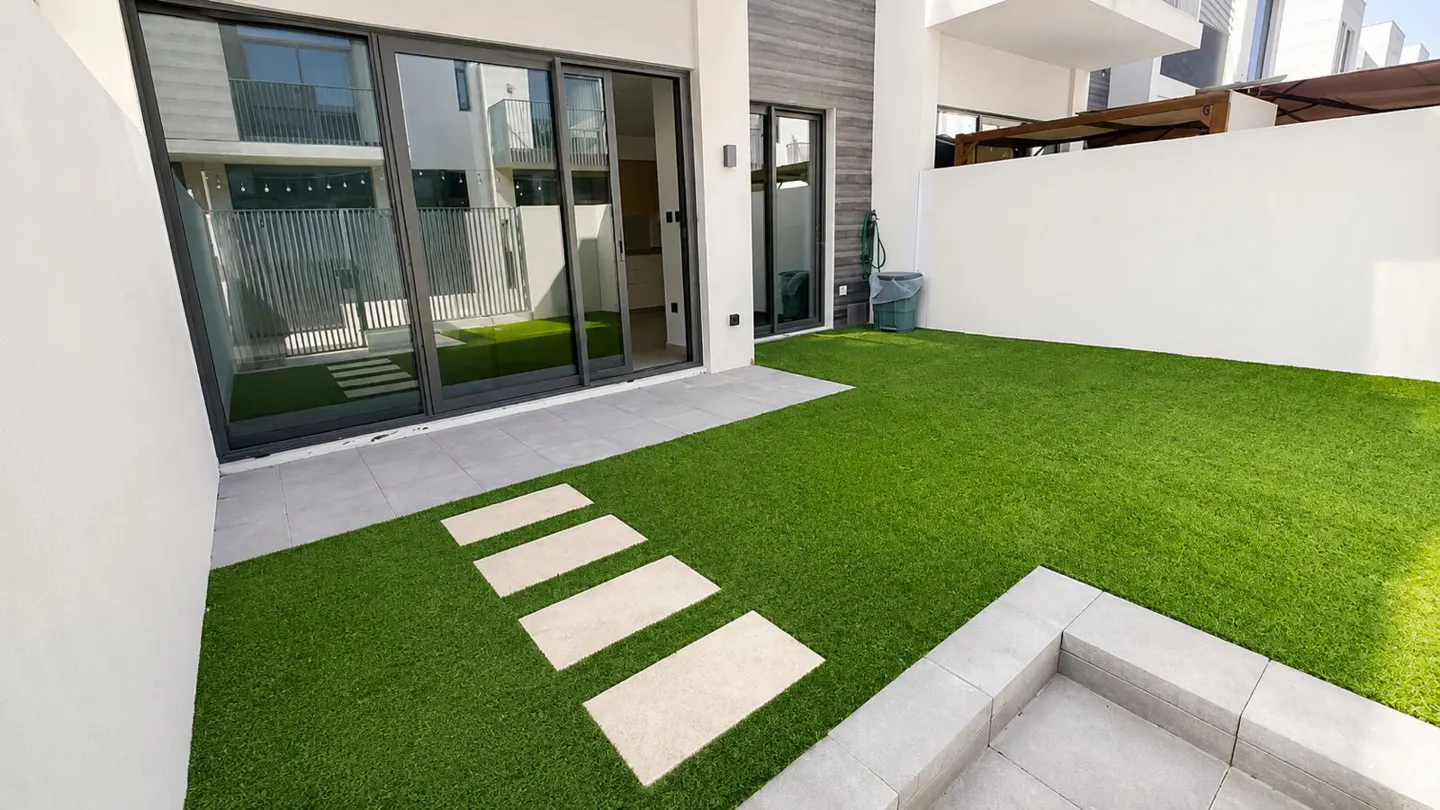 A modern home's backyard features green turf, stone pavers, and sliding glass doors. A white wall and a gray trash can are visible.