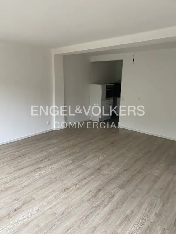 Empty room with light wood floors, white walls, and an open kitchen area. Engel & Völkers logo is visible.