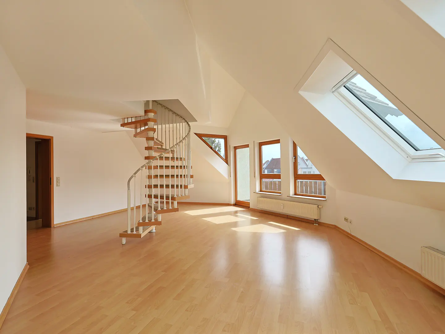Bright, empty attic apartment with wood floors, white walls, and a spiral staircase. Windows and a skylight let in natural light.