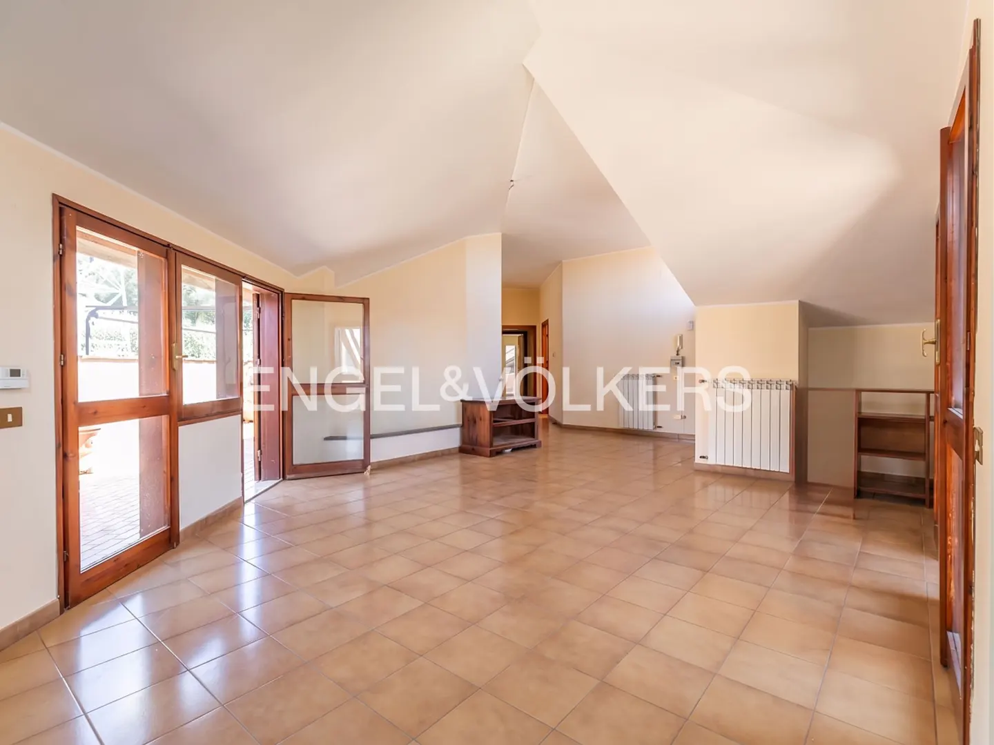 Spacious, empty room with beige walls, tiled floors, a vaulted ceiling, and wooden glass doors leading to a terrace.