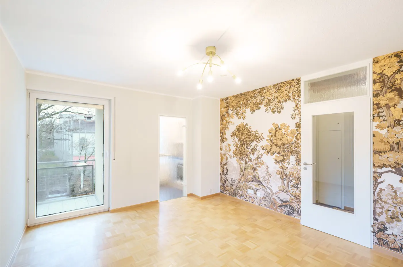 Bright, empty room with parquet floors, white walls, and a large window. One wall features a brown and white forest-themed wallpaper. A white door is on the right.