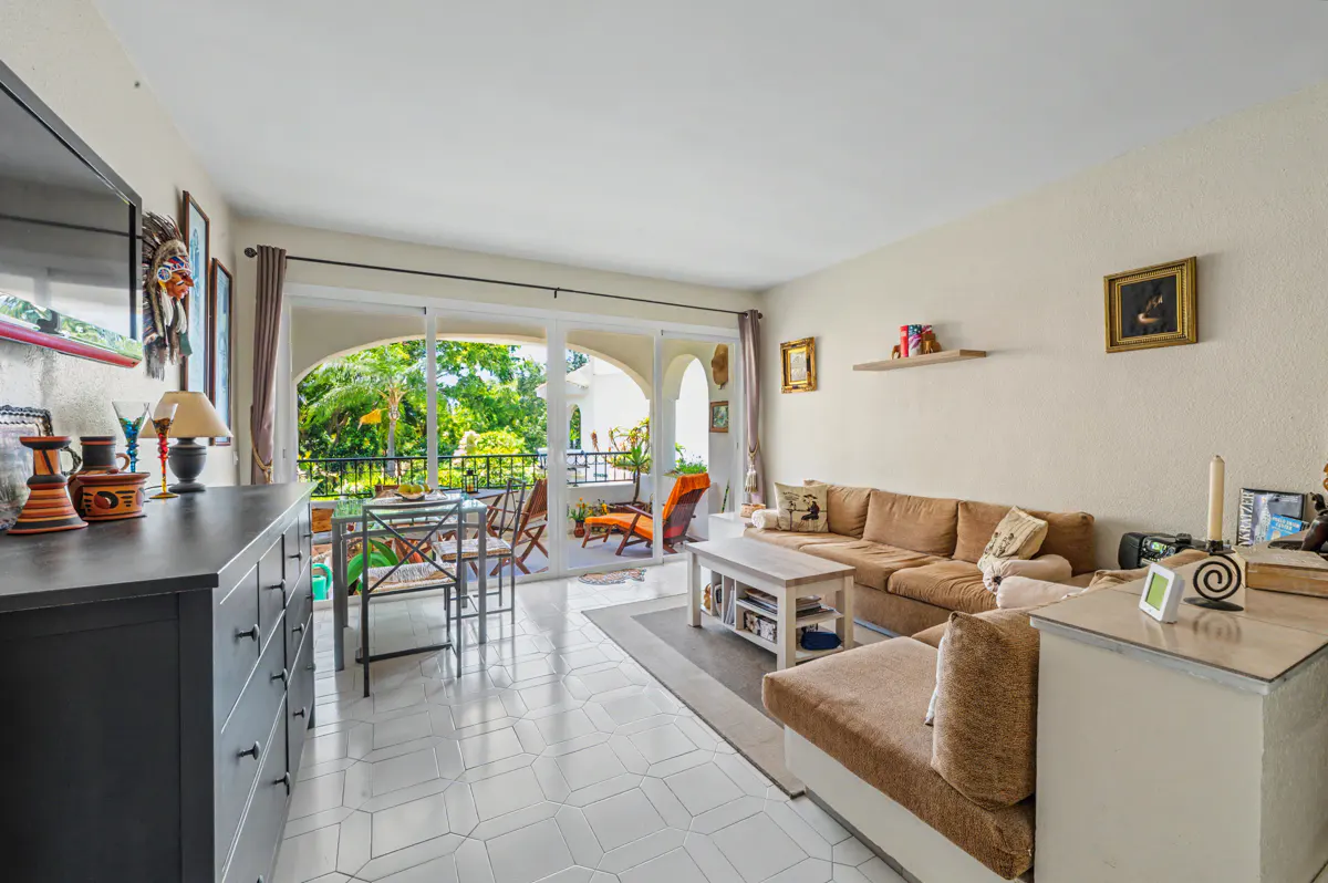 Bright living room with a brown sectional sofa, white tile floors, and a balcony with a table and chairs.