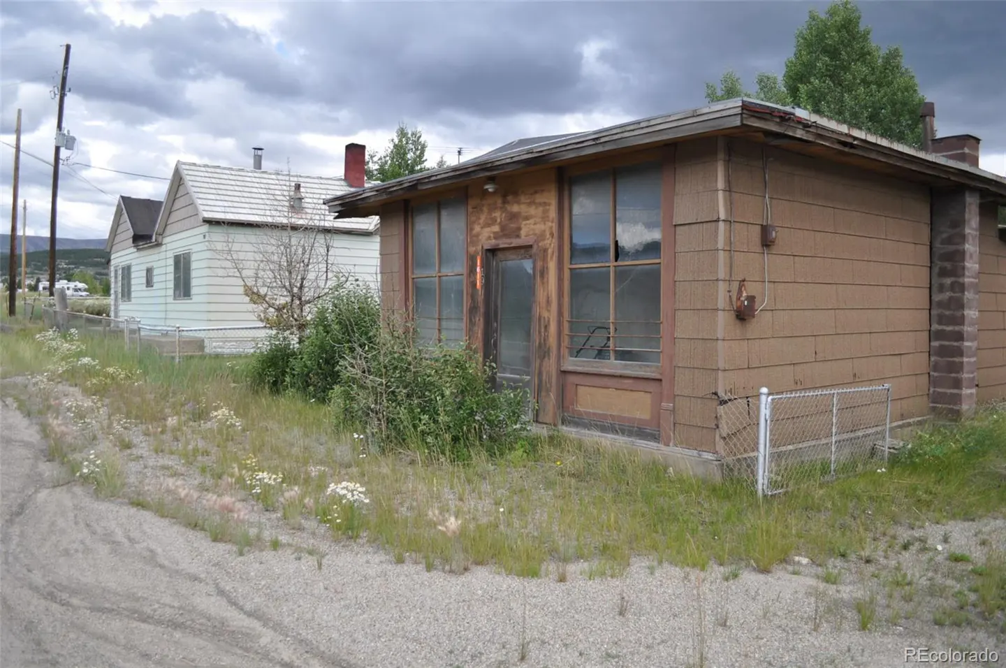 A weathered brown building with broken windows sits next to a white house under a cloudy sky.