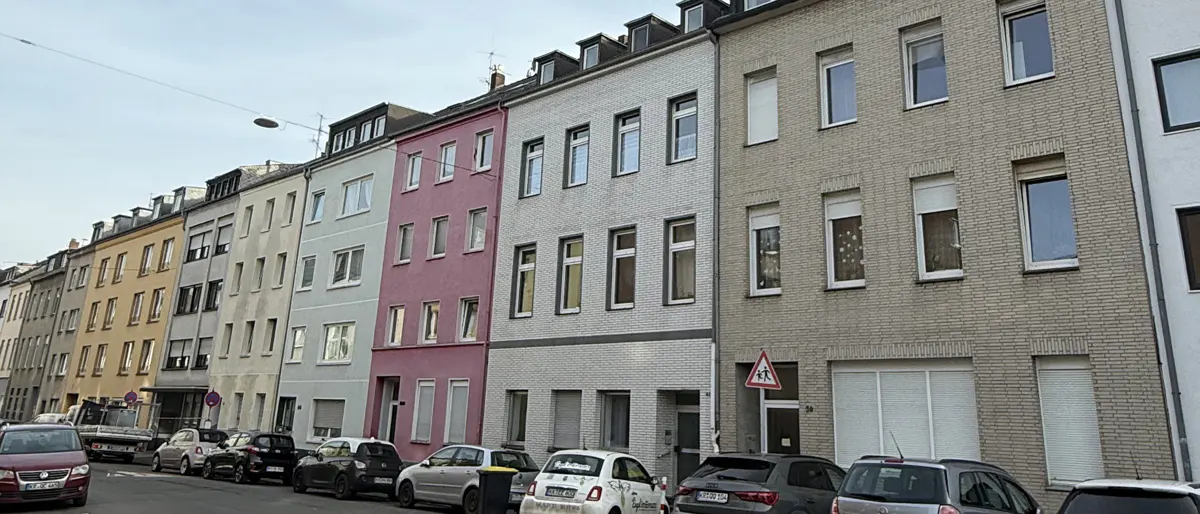 Street view of colorful, multi-story townhouses, cars parked along the curb, and a pedestrian crossing sign.