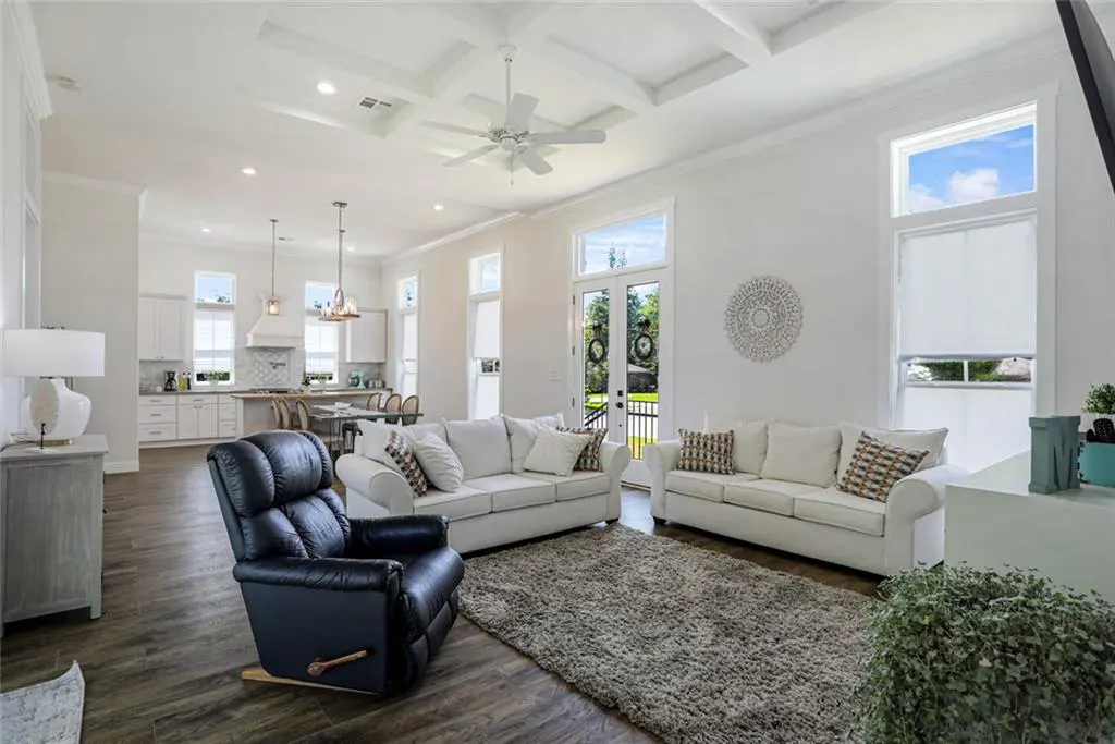 Bright, open-concept living room with white walls, dark wood floors, and a gray rug. Two white sofas face a black recliner. Kitchen visible in background.