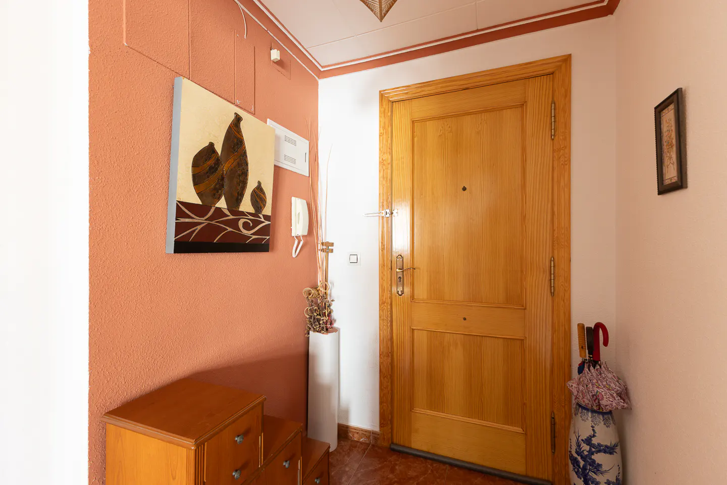 Entryway with a wooden door, terracotta walls, and a painting of vases. A wooden dresser sits below the painting. An umbrella stand is near the door.