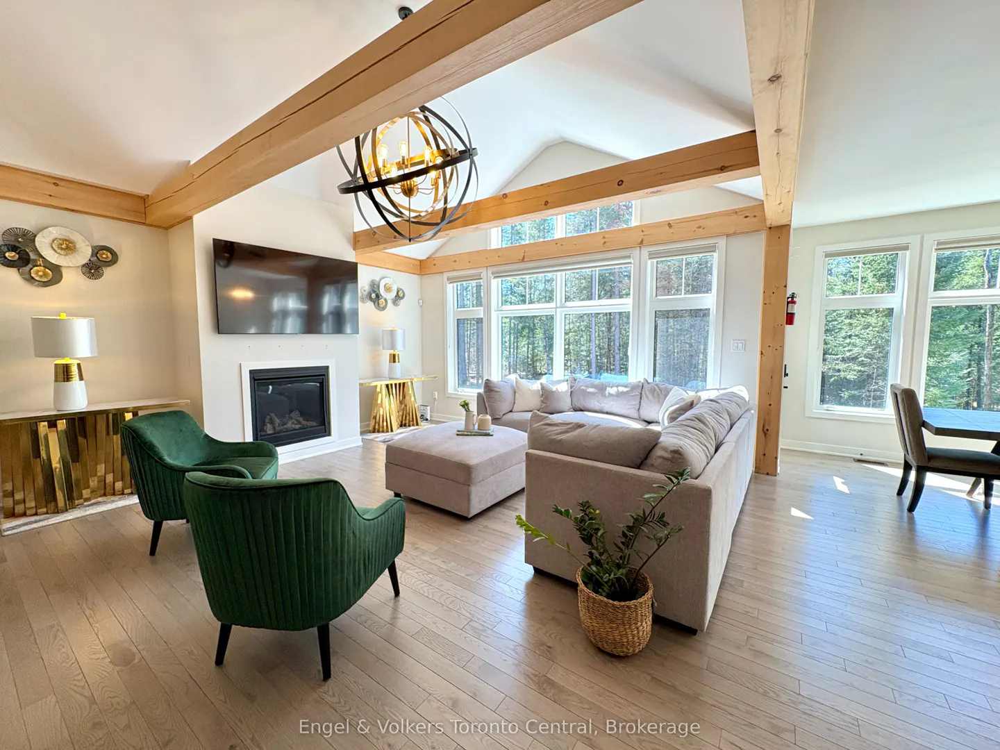 Bright living room with wood floors, beams, and large windows. Green chairs face a fireplace with a TV above. A beige sectional sofa sits near the windows.