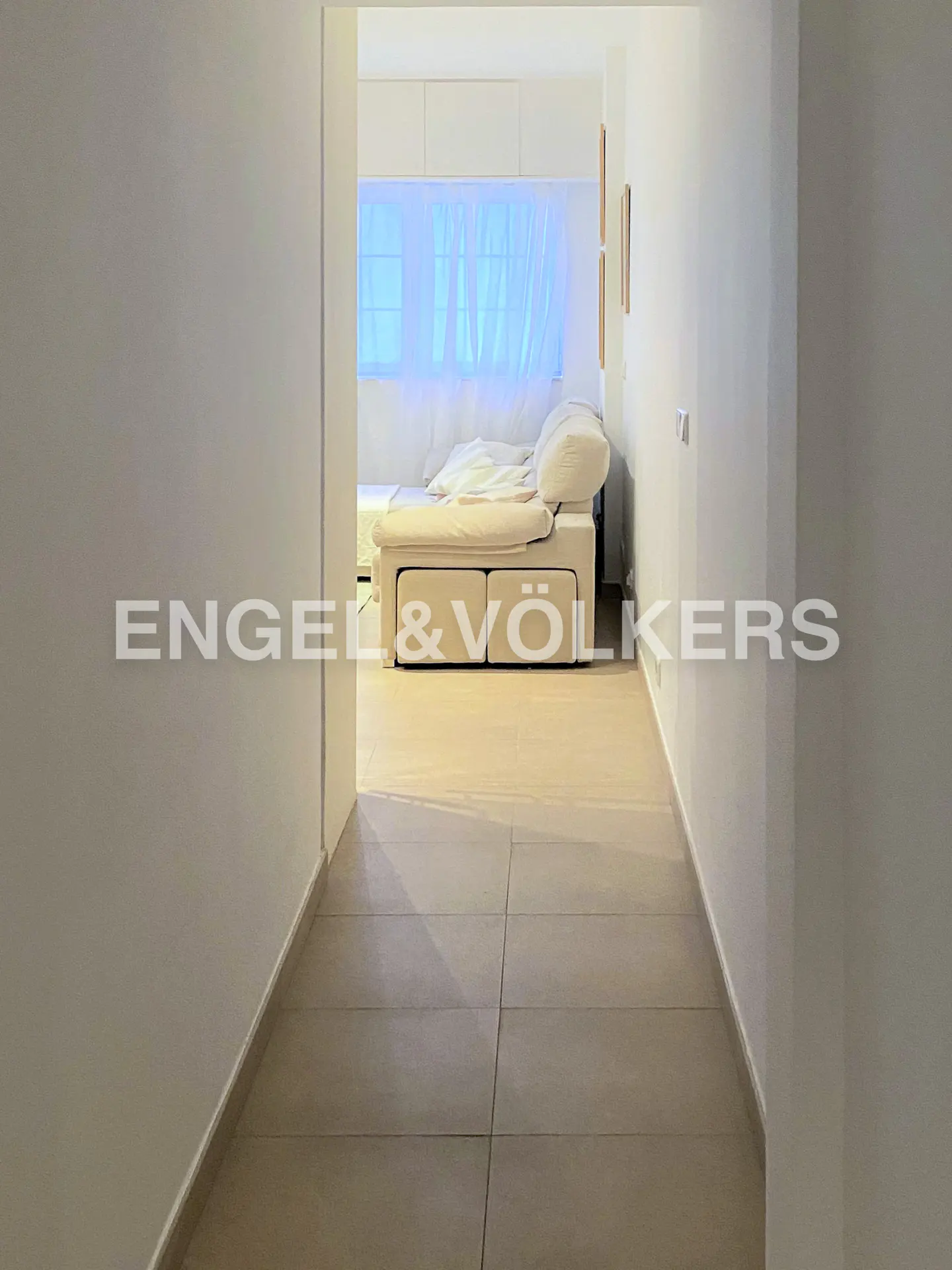 Hallway view into a bright room with a beige sofa and white walls. The floor is tiled. Engel & Völkers logo is visible.
