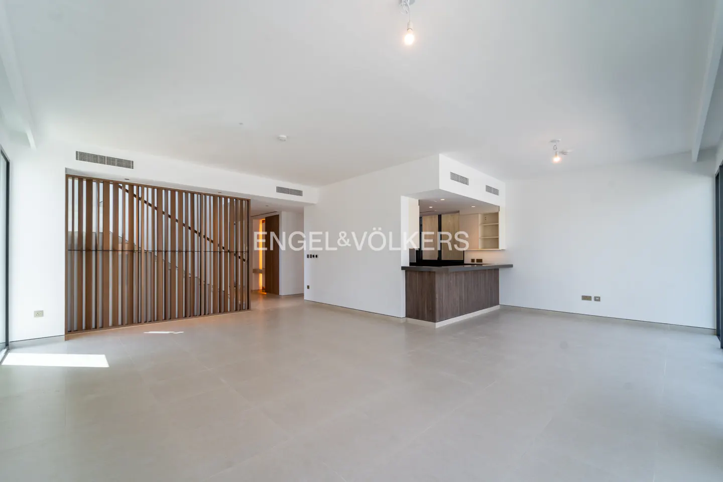 Modern open-plan living area with white walls, light tile floors, a wooden slat room divider, and a dark wood kitchen island.