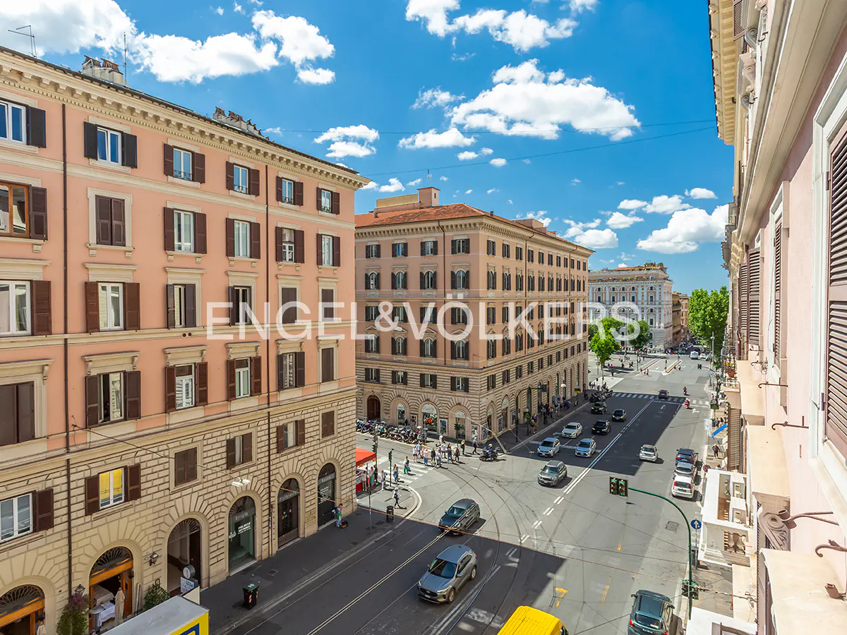 View of a busy street in Rome, Italy, with pink buildings, cars, and pedestrians under a blue sky with white clouds.