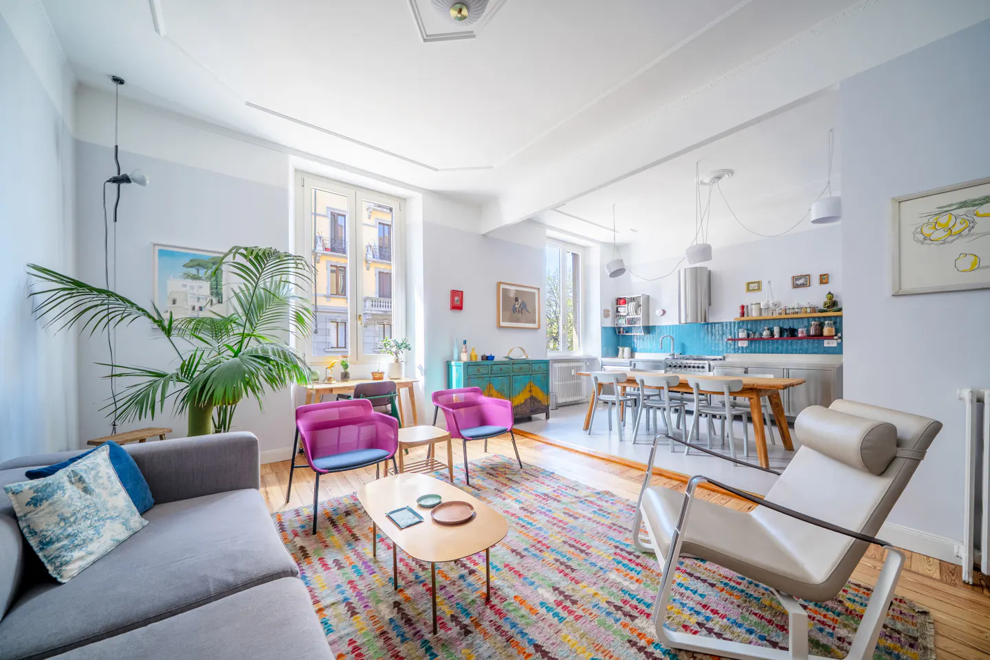 Bright, open-concept living space with a gray sofa, pink chairs, and a colorful rug. Kitchen with blue backsplash in the background.