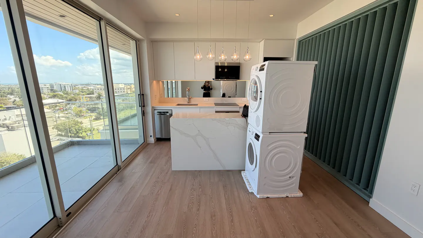 Bright, modern kitchen with white cabinets, island, and stacked washer/dryer. Large sliding doors open to a balcony with city views.