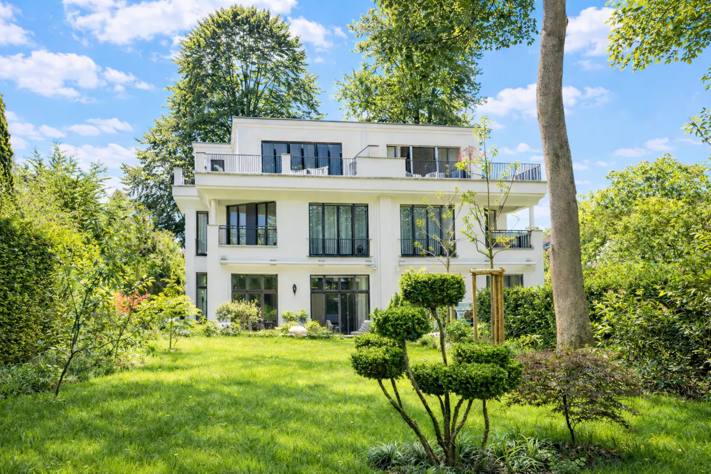 A white three-story modern villa with black-framed windows and balconies, set in a lush green garden under a blue sky.