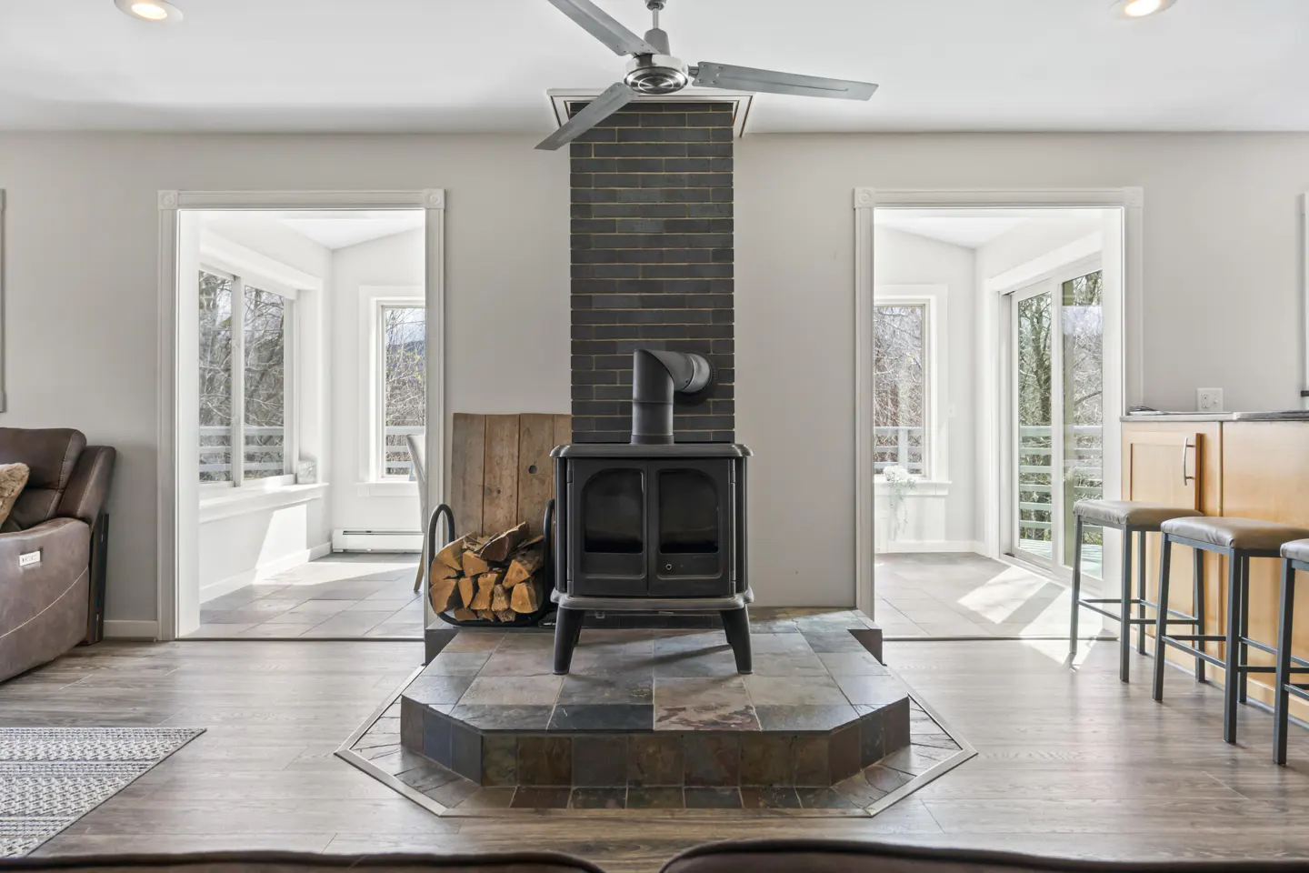 Living room with a black wood-burning stove and brick chimney. Wood is stacked next to the stove. Two doorways lead to sunrooms.
