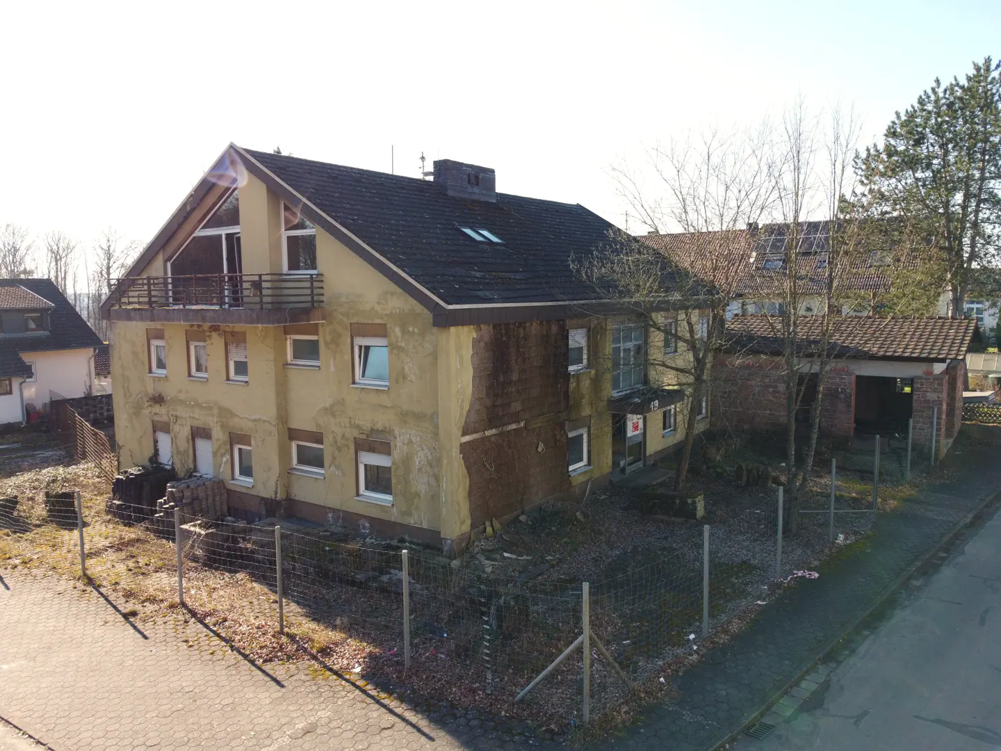Exterior view of a two-story yellow house with a dark roof, a balcony, and a fenced yard.