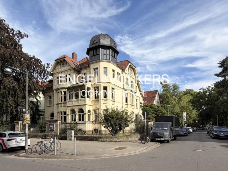 A historic yellow villa with a dark turret stands on a street corner with parked cars, bicycles, and lush green trees. [AI-generated text]