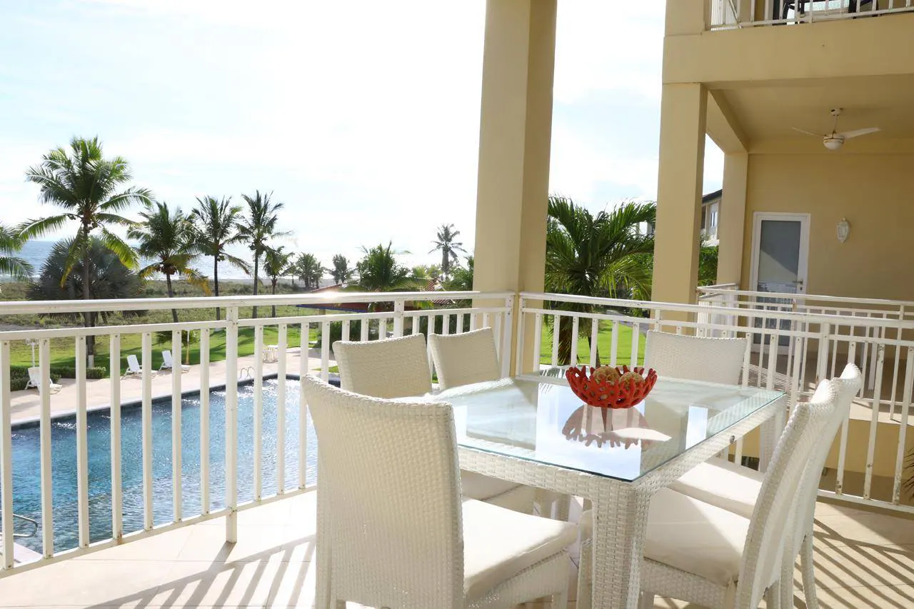 Balcony view with white wicker table and chairs, overlooking a pool, palm trees, and the ocean.