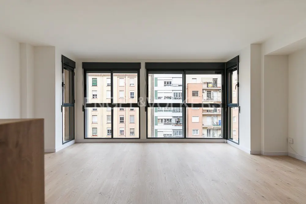 Empty room with light wood floors and large black-framed windows overlooking urban apartment buildings.
