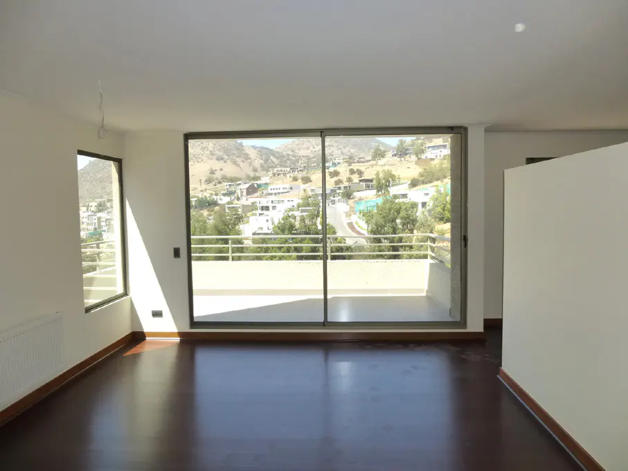 An empty room with dark wood floors and white walls. A large sliding glass door leads to a balcony with a view of a hillside neighborhood.