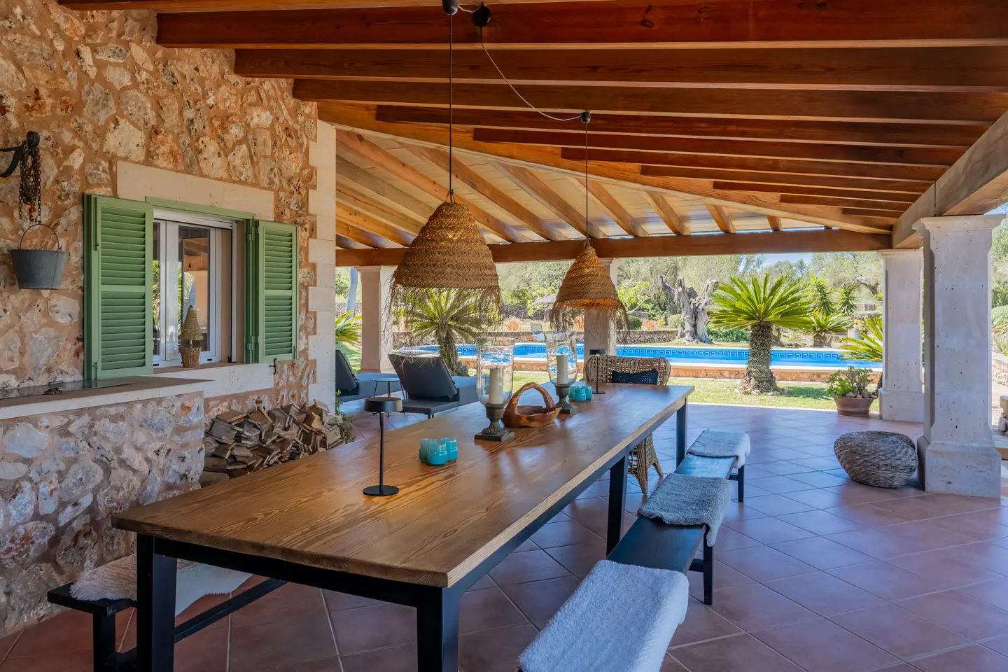 Covered patio with a long wooden table, benches, and straw pendant lights. A pool and palm trees are visible in the background.