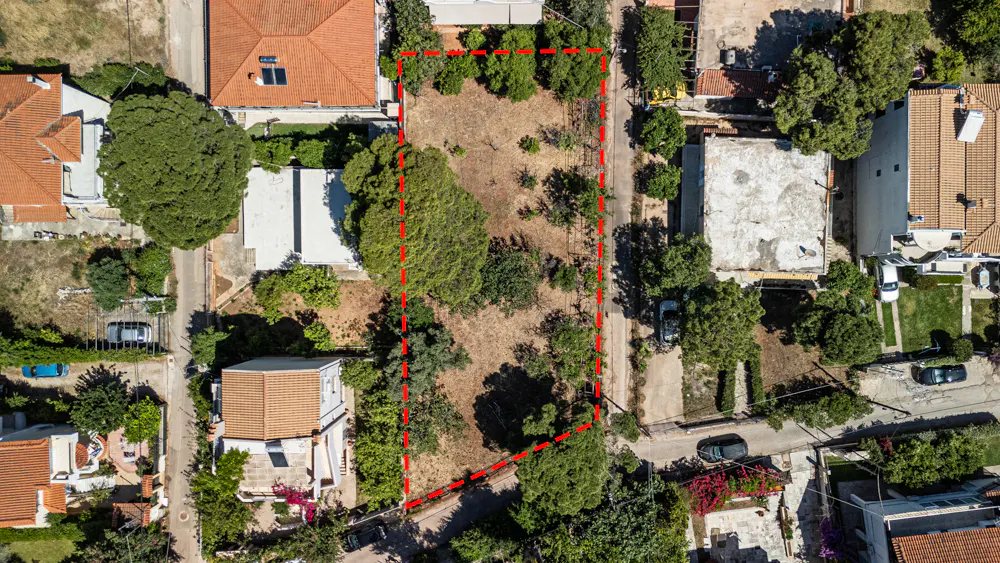 Aerial view of a vacant rectangular plot of land outlined in red, situated between residential houses and trees.