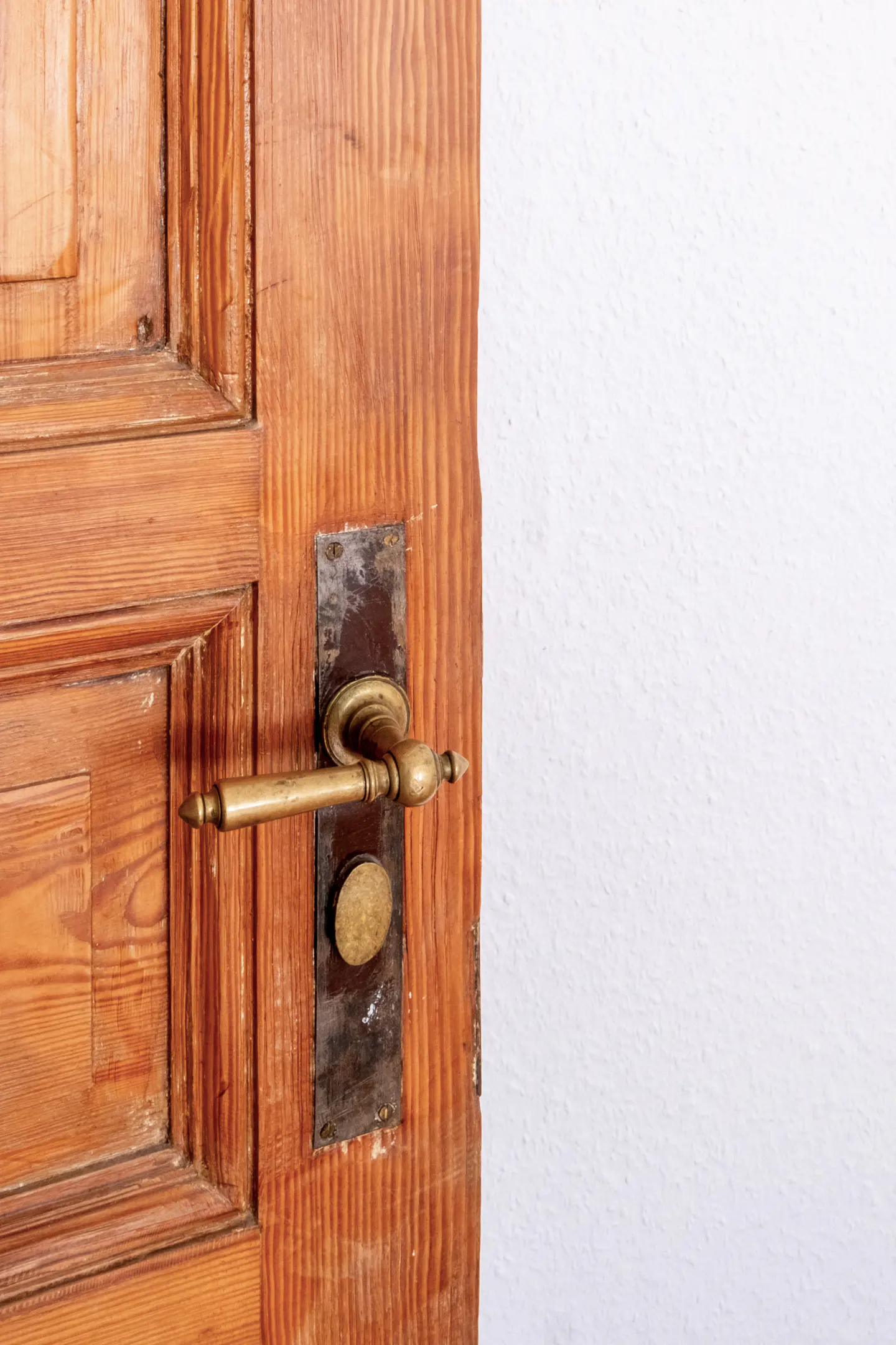 Close-up of a partially open, paneled wooden door with a brass handle and a white wall in the background.