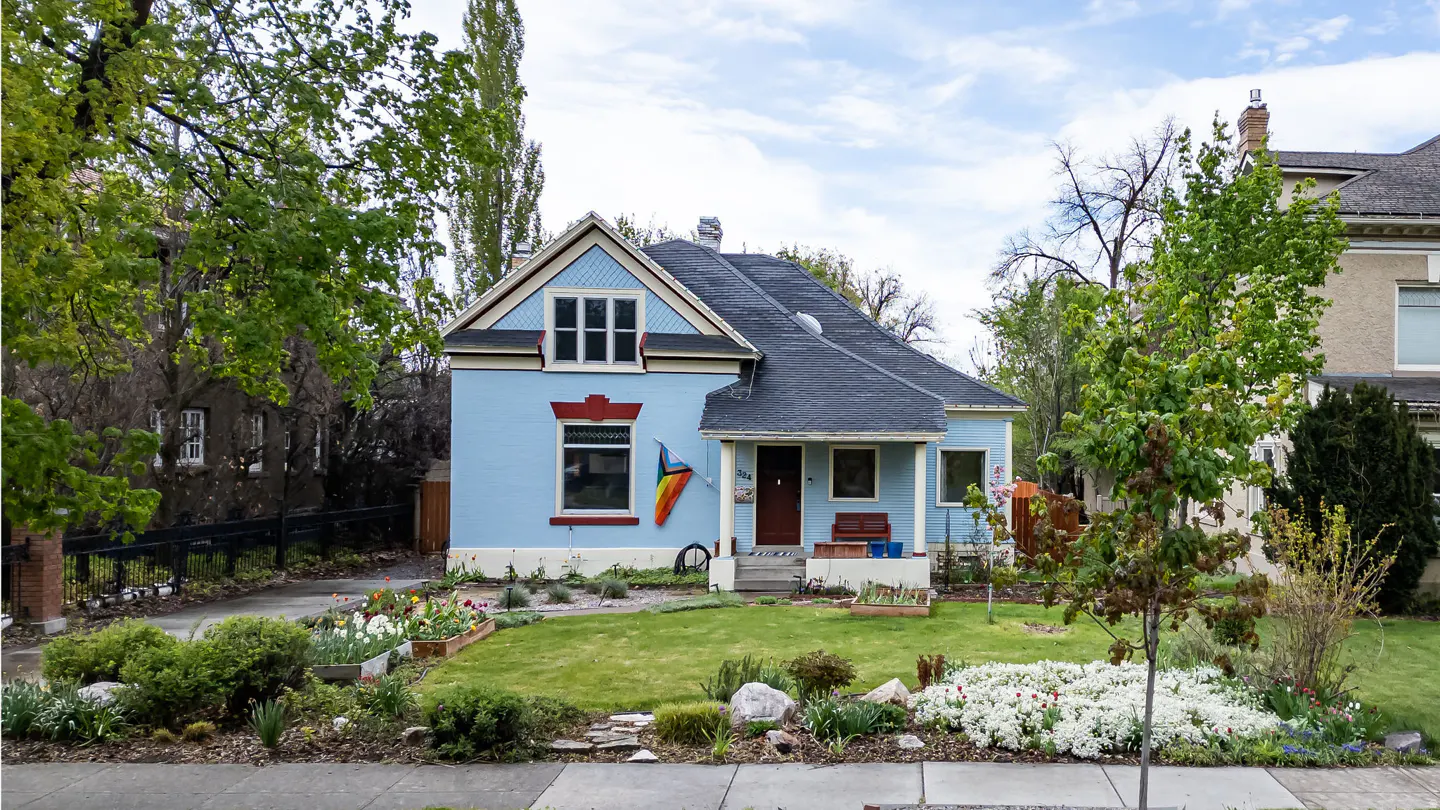 Light blue house with a dark roof, a rainbow flag, and a green lawn with flowerbeds.