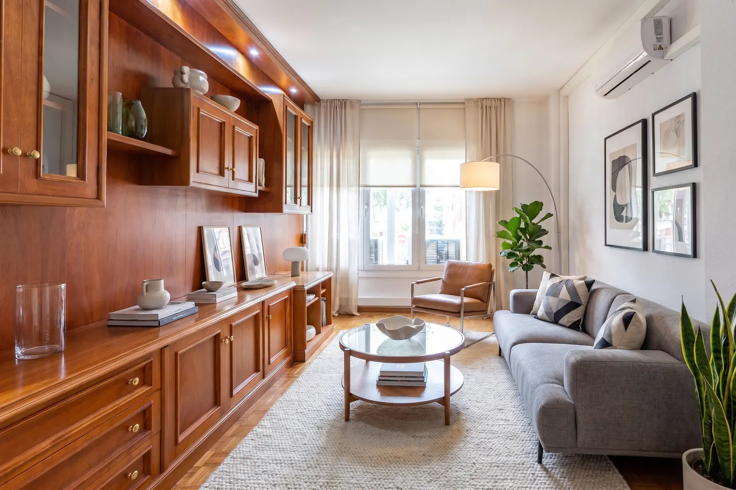 Living room with wood cabinetry, grey sofa, glass coffee table, leather armchair, and large window with light curtains.
