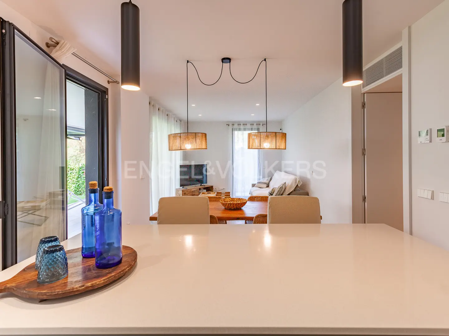 A bright, modern interior with a white countertop, blue bottles, and a dining area with a wooden table and chairs. Cylindrical pendant lights hang above.