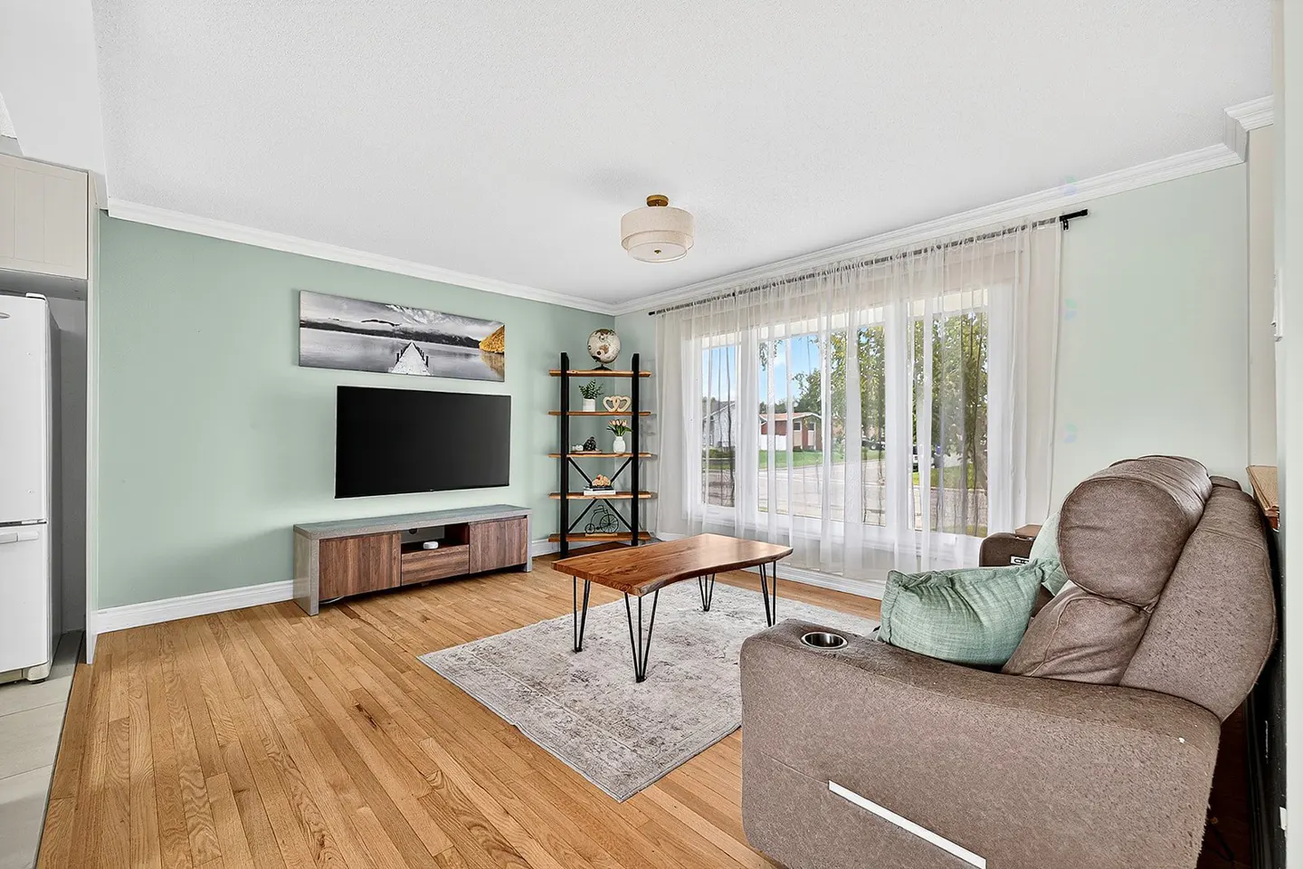 Living room with wood floors, light green walls, and large windows with sheer curtains. A brown recliner sits on a patterned rug near a wood coffee table. A TV and bookshelf are against the wall.