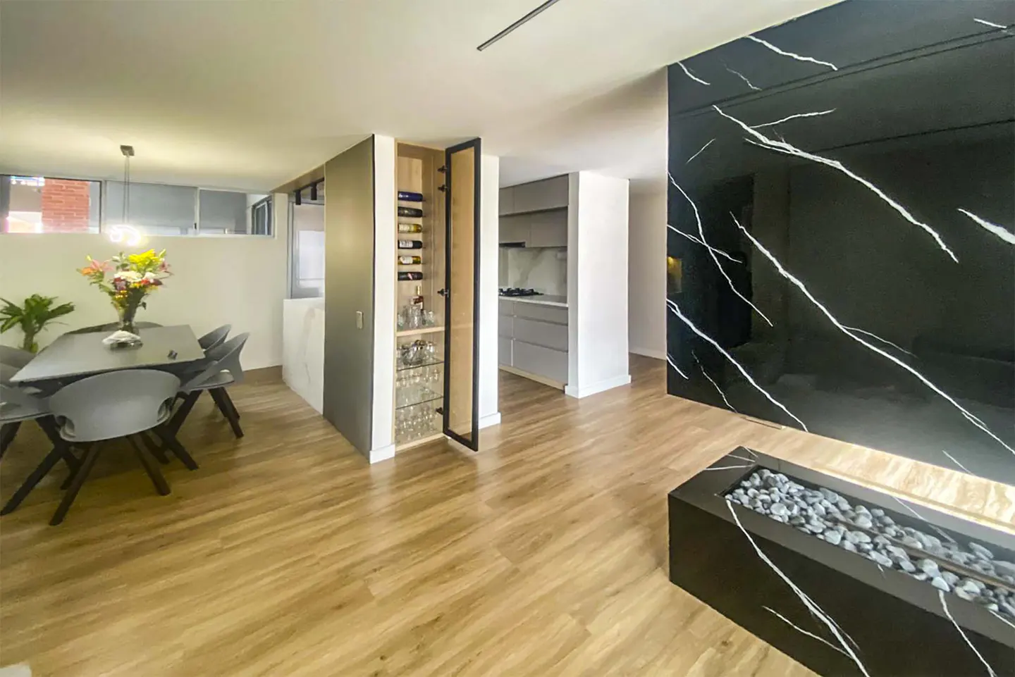 A modern apartment interior with a dining area, wine cabinet, kitchen, and black marble accent wall with a fireplace.