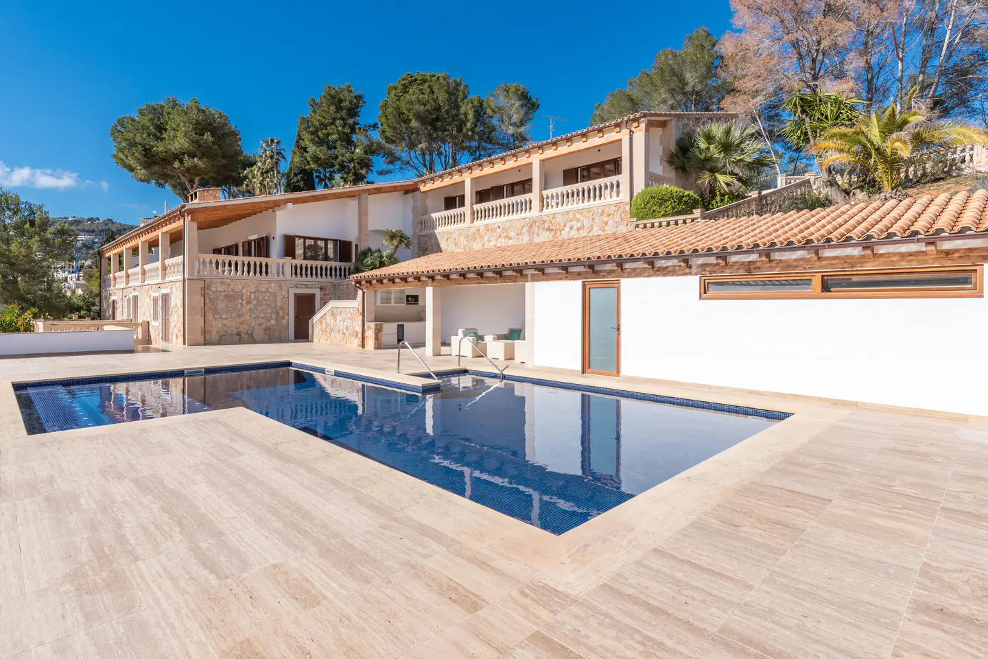 A luxurious villa with a blue tiled pool and beige stone patio under a clear blue sky.
