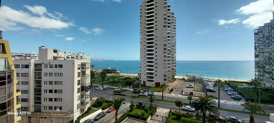 View of a city beach with tall buildings, palm trees, cars, and blue ocean under a partly cloudy sky.