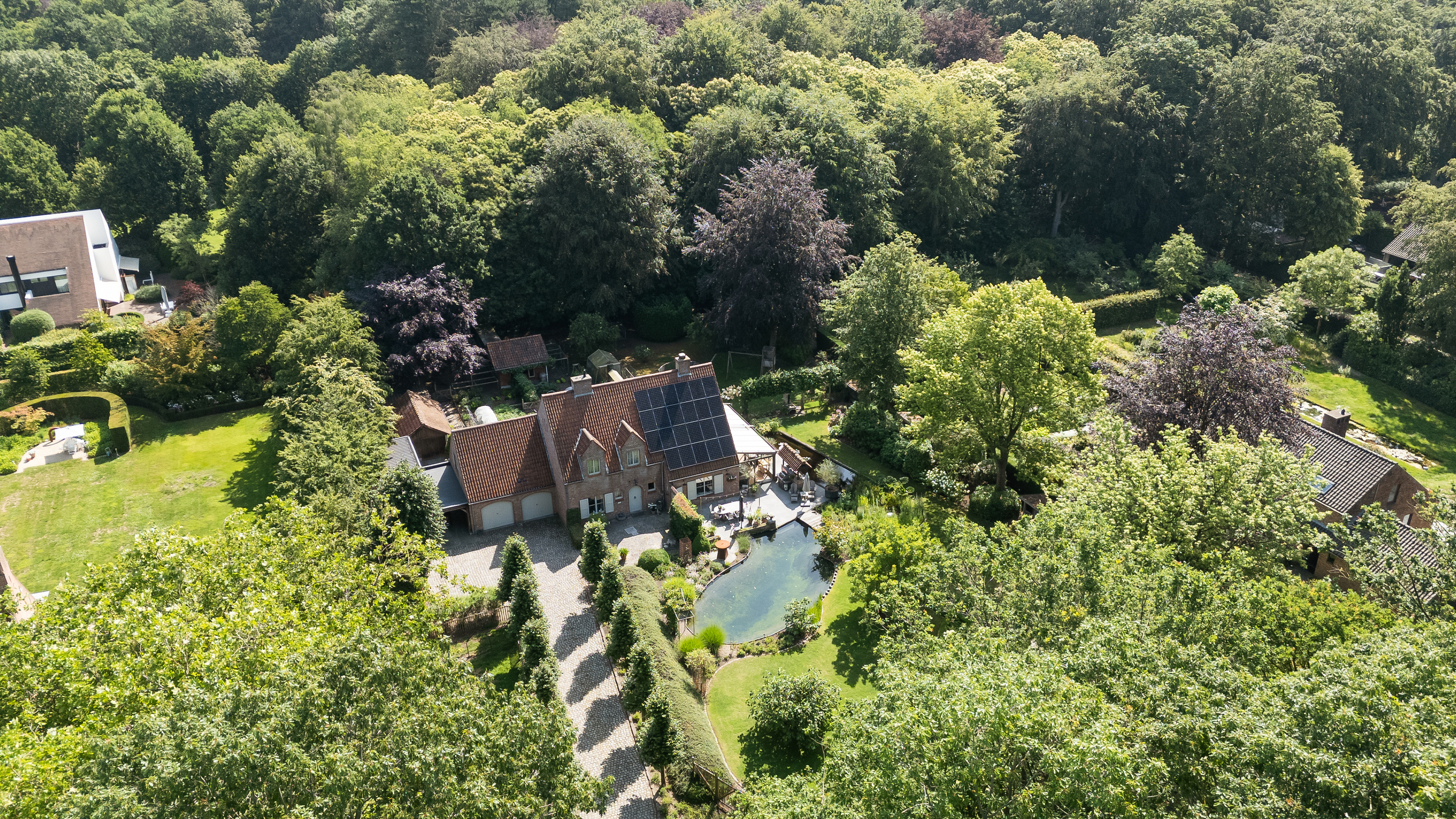 Aerial view of a brick house with solar panels, a driveway, and a pond, surrounded by lush green trees and gardens.