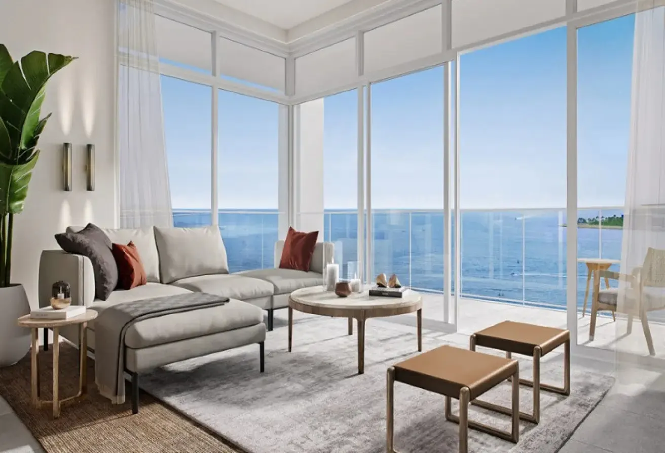 Bright living room with a gray sectional sofa, round coffee table, and floor-to-ceiling windows overlooking the ocean.