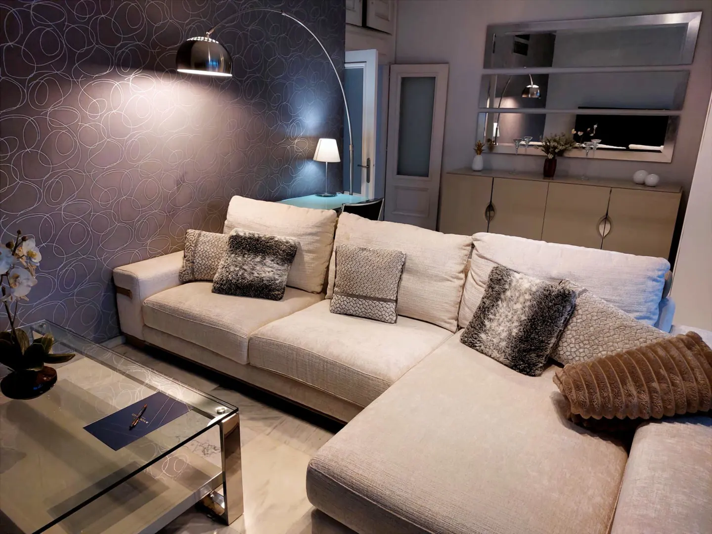 Living room with a beige sectional sofa, patterned wallpaper, and a glass coffee table. A floor lamp arches over the sofa.