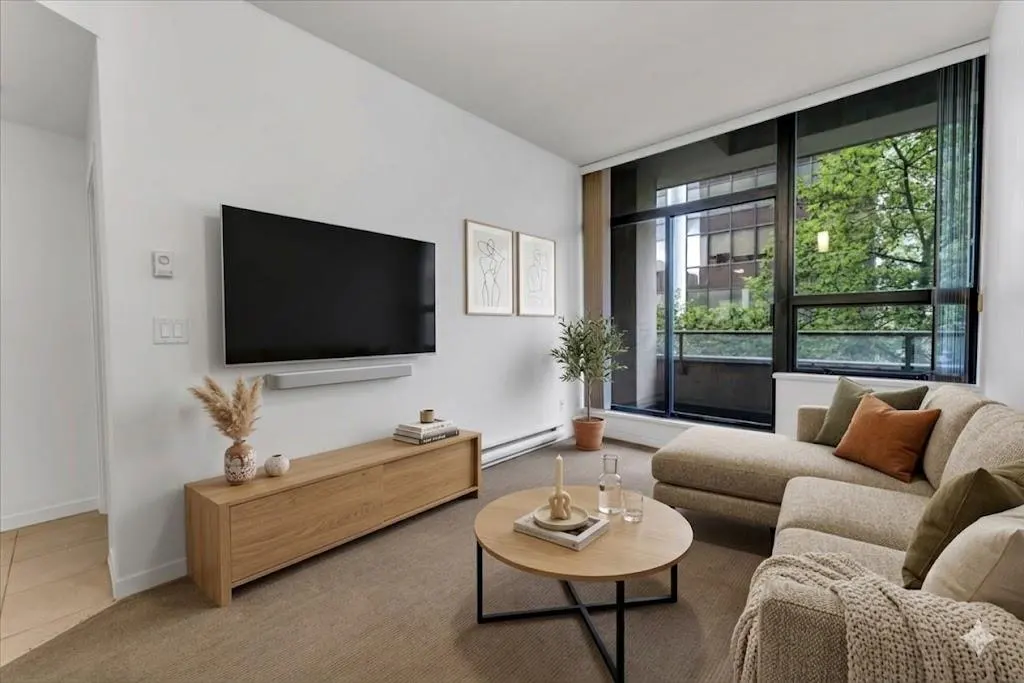 Living room with a beige sectional sofa, round coffee table, and a large window with a view of trees. A TV is mounted on the white wall.